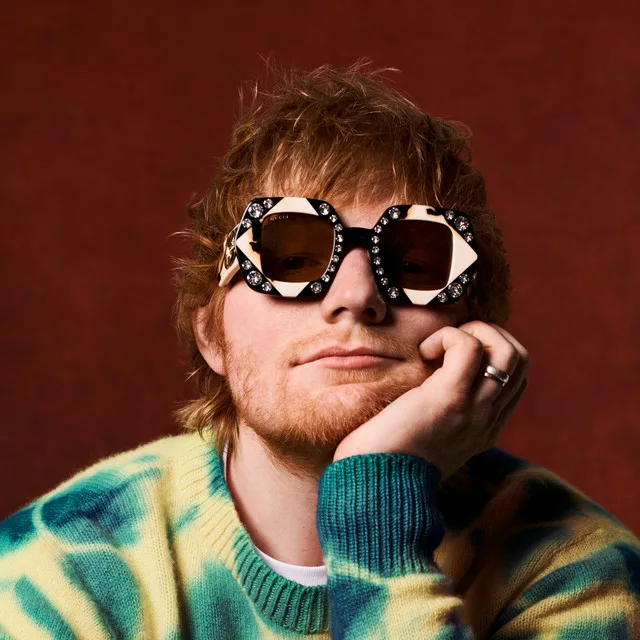 Ed Sheeran smiling, holding an acoustic guitar, and surrounded by festive Christmas lights