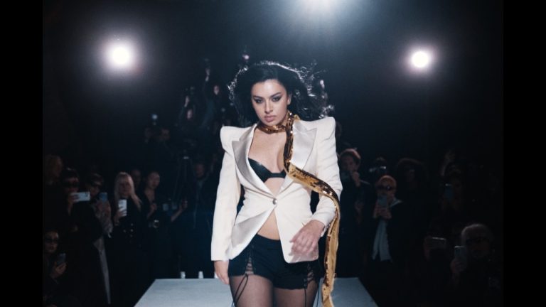 Charli XCX strutting down a dark runway lit by harsh spotlights in the official SS26 music video.